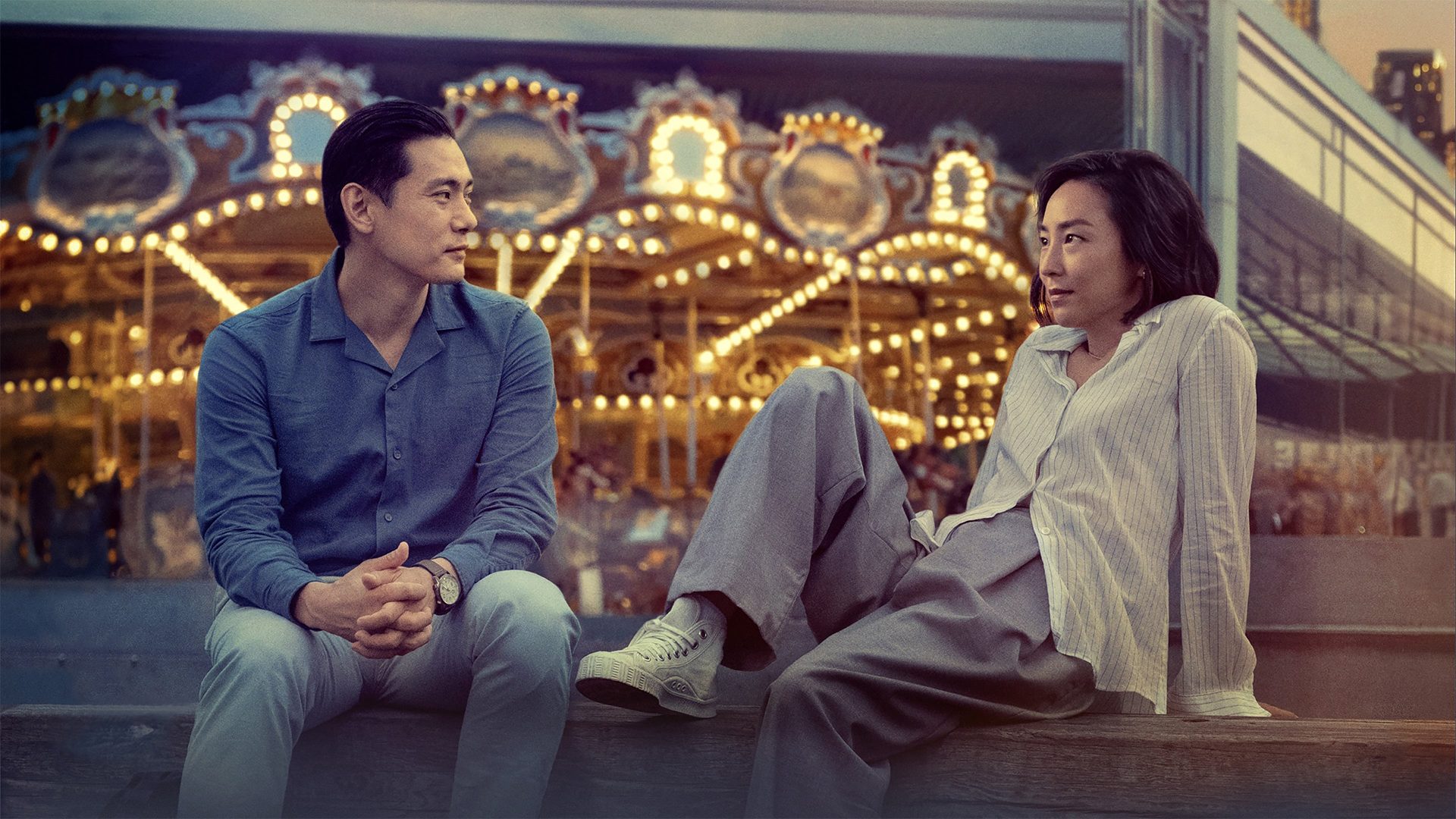 Nora and Hae Sung sit in front of a carousel by Brooklyn Bridge, New York City.